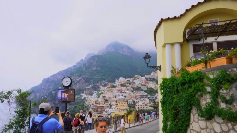 Tourists walking and taking photos in Positano with colorful hillside houses and mountains in the background